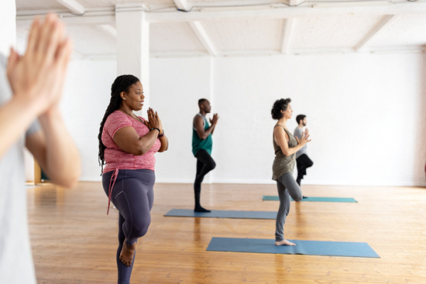 A group of people doing a standing pose in a yoga class; a woman wearing a pink top and dark purple leggings in the foreground along with a blurred paire of hands